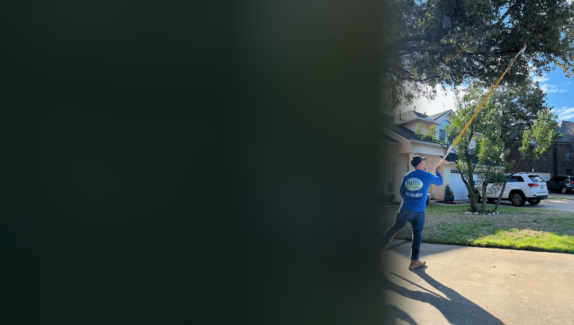 JW crew member in a blue company shirt pole pruning a large oak over a driveway while a coworker assists